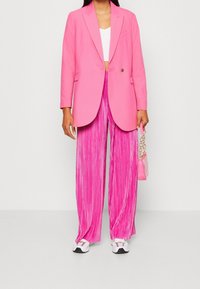 Světle růžový blazer s vypasovaným střihem, kombinovaný s širokými plisovanými kalhotami ve stejné barvě. Doplněný malou růžovou kabelkou.