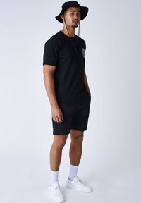 Camiseta negra con diseño gráfico, pantalones cortos negros, zapatillas blancas y un sombrero tipo bucket negro. Tiene un ajuste atlético y un estilo casual.