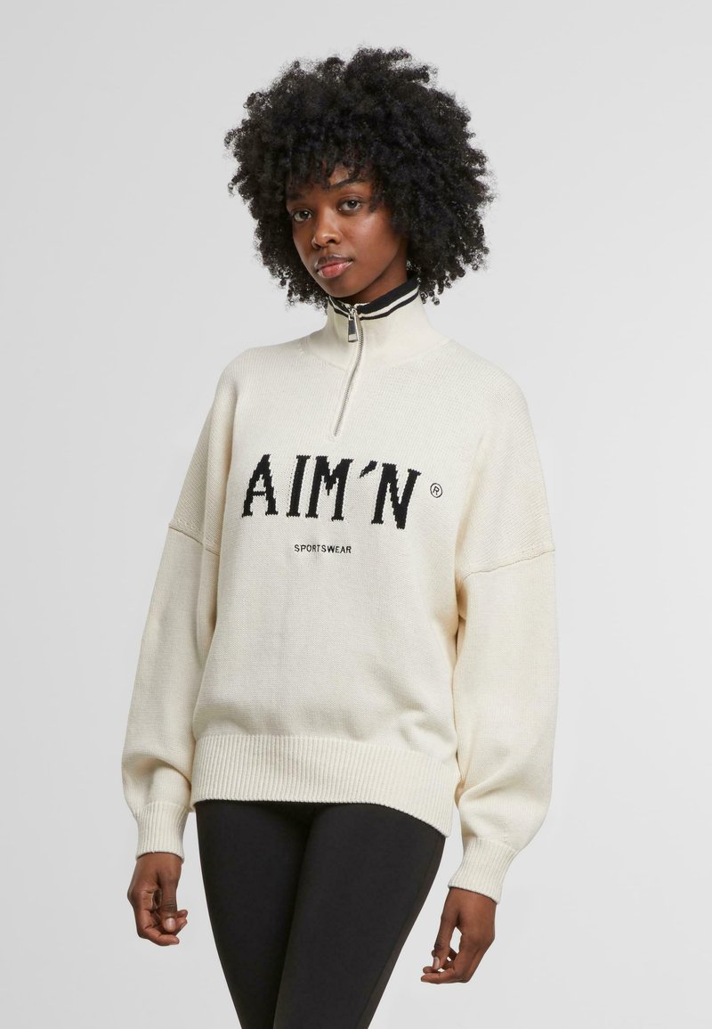 Cream-colored sweater with a high collar and half zip, featuring a black logo "AIM'N" in bold letters across the front. Ribbed cuffs and hem.