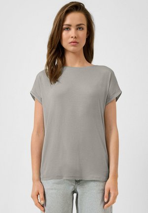 Jonge vrouw met losbruin haar, gekleed in een effen grijs T-shirt met korte mouwen en lichte spijkerbroek, staand tegen een witte achtergrond.