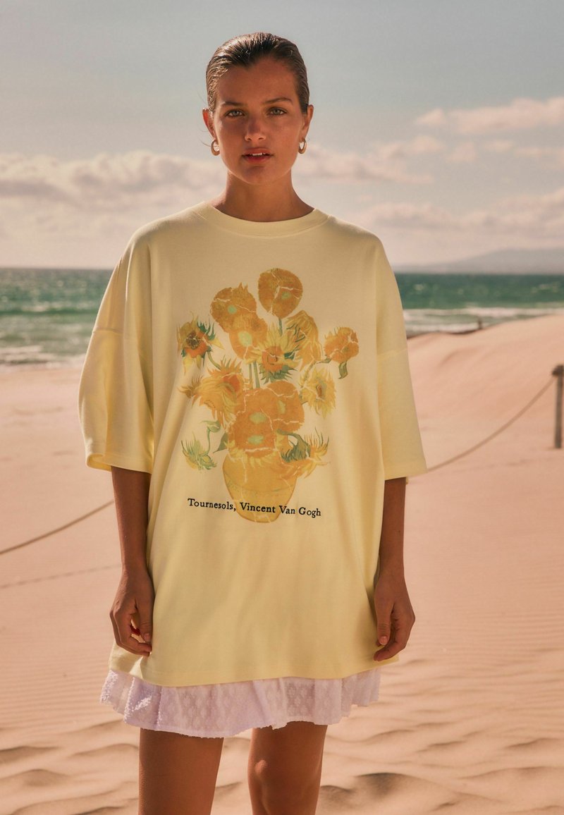 Mujer de pie en una playa de arena, vestida con una camisa amarilla de gran tamaño con la pintura de girasoles de Vincent Van Gogh y una falda blanca.