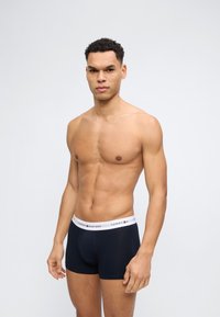 Muskelöser Mann mit kurzen lockigen Haaren, der marineblaue Tommy Hilfiger Boxershorts trägt und vor einem schlichten hellgrauen Hintergrund steht.