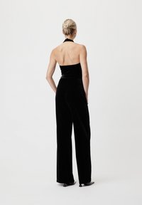 Svart fløyels jumpsuit med halterneck-design, vid benåpning og subtil glans, vist fra ryggen. Enkel og elegant stil.