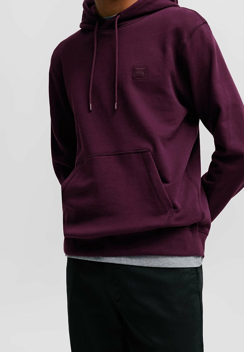Bordeauxfärgad hoodie tillverkad av mjukt tyg, med en framficka, dragsnören och en liten präglad logotyp på bröstet.