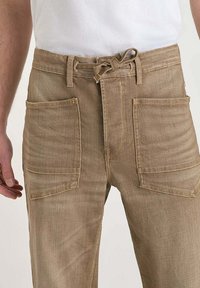 Un homme porte un pantalon beige clair avec de grandes poches avant à rabas et une ceinture en tissu nouée, associé à une chemise blanche.