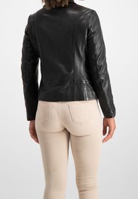 Černá kožená bunda s přiléhavým střihem, hladkou texturou a detailem na zádovém švu, v kombinaci se světle béžovými kalhotami.