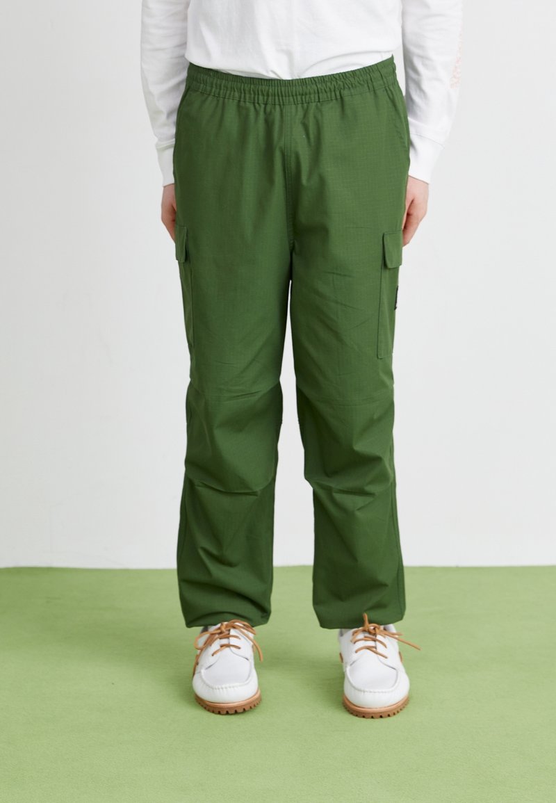 Gröna cargo-byxor med elastiskt midjeband, med sidofickor och en slät textur, tillsammans med vita skor med bruna sulor.