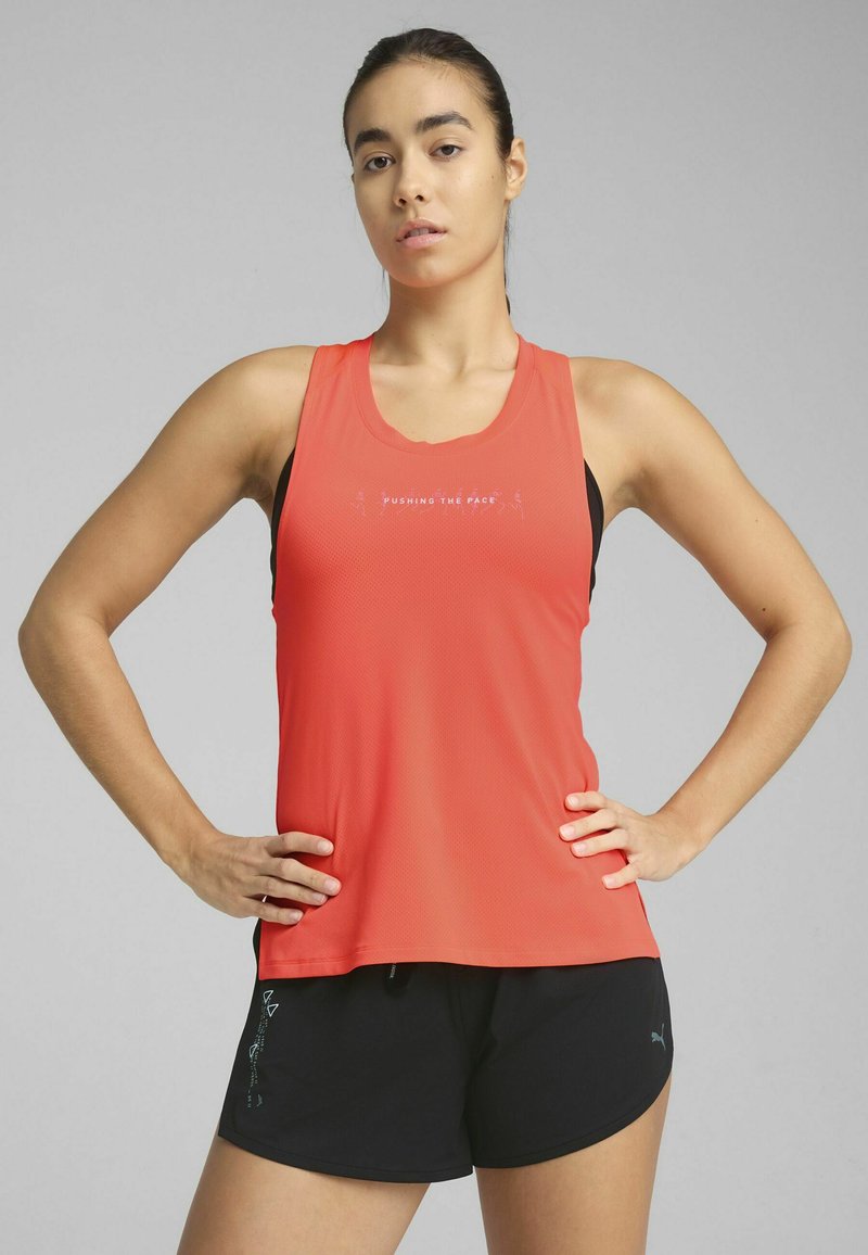 Punane sportlik tank top, millel on võrgustruktuur ja graafika "PUSHING THE PACE"; koos mustade lühikeste pükstega, millel on väike logo ja minimaalne disain.