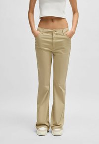 Beige, flared trousers with a smooth texture, front pockets, a button closure, and contrast stitching. Paired with white sneakers.