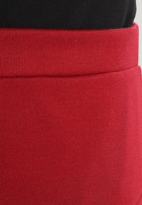 Röd stretchig tyg med en slät yta, med en bred midjeband och en svart tygsektion ovanför. Sömlös design utan synlig hårdvara.