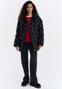 Czarna pikowana kurtka puchowa z różowym kwiatowym haftem, srebrnym zamkiem, wysokim kołnierzem; nałożona na czerwoną bluzę i zestawiona z czarnymi dżinsami i butami.