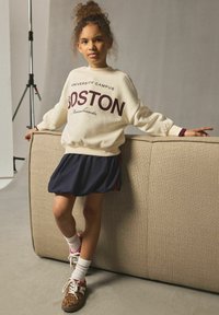 Krämfärgad sweatshirt med texten "BOSTON", i kombination med en mörkblå kjol. Bruna sneakers med leopardtryck och rosa snören samt vita crewstrumpor.