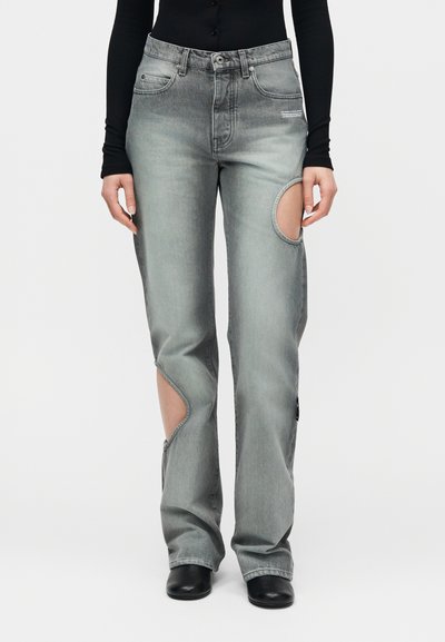 Γκρι ξεβαμμένο τζιν με μεγάλα κυκλικά κοψίματα στο δεξί μηρό και το αριστερό κάτω πόδι, φορεμένο με μαύρο τοπ και μαύρα παπούτσια.