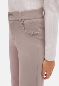 Pantalones elásticos de color rosa claro con detalle de bolsillo con volantes y botón metálico en forma de corazón. Tejido mezclado de algodón, diseño ajustado.