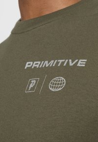 Camiseta de algodón verde oliva con el texto bordado en blanco "PRIMITIVE" y dos símbolos: un ícono y un globo. Textura suave, corte clásico.