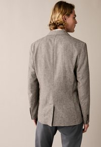 Grauer Blazer aus strukturiertem Stoff, mit einem Schlitzt auf der Rückseite und Knöpfen an den Manschetten. Kombiniert mit maßgeschneiderten grauen Hosen.