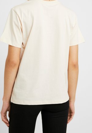 Persoon met een effen gebroken witte korte mouw t-shirt gezien van achteren, gecombineerd met zwarte strakke broek tegen een neutrale achtergrond.