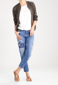 Giacca bomber verde oliva scuro, t-shirt bianca, jeans blu strappati con ricami floreali e sandali marroni con cinturini, posato in modo informale.