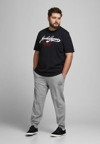 Camiseta negra con logo blanco y texto rojo, combinada con pantalones de chándal grises que tienen un logo redondo en el muslo, completado con zapatillas negras.