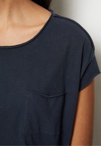 Ingrandimento di una persona che indossa una maglietta a maniche corte di colore blu scuro con taschino sul petto, che mostra il collo e la spalla.