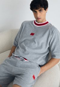 Graues Sportset mit einem kurzärmligen Oberteil und einem rot-weißen Kragen sowie passenden Shorts. Enthält ein Marken-Patch auf der Vorderseite.