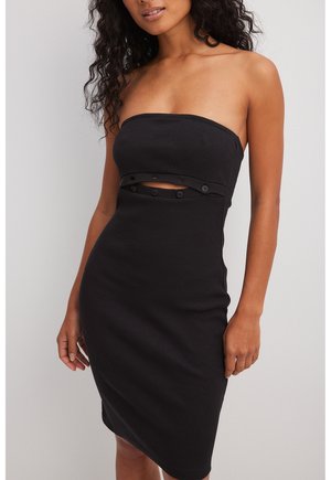 Femme portant une robe midi noire sans bretelles, côtelée, avec une découpe frontale fixée par des boutons.