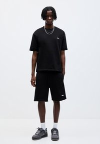 Sort bomulds t-shirt med rund hals, parret med matchende sorte shorts, begge med diskrete logo detaljer. Båret med mørke sneakers og hvide sokker.