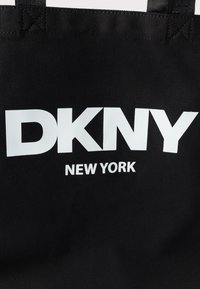 Saco tote preto feito de tecido resistente, com texto branco "DKNY NEW YORK" exibido de forma destacada em fonte negrito.