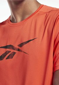 Camiseta atlética de color naranja brillante con cuello redondo, que presenta un diseño de logo en negro. La tela parece ligera y suave al tacto.