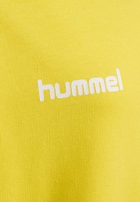 Gult tyg med en jämn textur som har den vita logotypen "hummel" tydligt visad. Designen är enkel och modern.