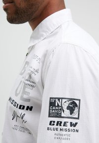 White long-sleeve shirt featuring various printed texts and graphics on the front and sleeve, with a structured collar and lightweight fabric.