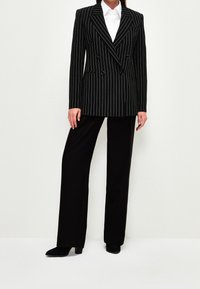 Nainen, joka on pukeutunut mustaan pinstripe kaksiriviseen bleiseriin, valkoiseen kauluspaitaan, mustiin leveälahkeisiin housuihin ja mustiin korkokenkiin, seisoo tasaisella taustalla.