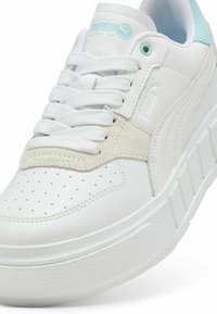 Fehér sportcipők sima bőr felsőrésszel, perforált orral, szürke velúr díszítéssel és fehér platformtalpával. Jellemzője a türkiz logó és a fűzőlyuk.