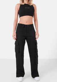Crop top noir côtelé avec logo, associé à un pantalon cargo noir doté de multiples poches et d'une coupe droite. Des baskets blanches complètent la tenue.