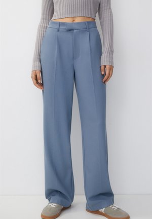 Person, die hochtaillierte, weit geschnittene blaue Hose, grauen, kurz geschnittenen, gerippten Pullover und graue Turnschuhe trägt und vor einem hellen Hintergrund steht.