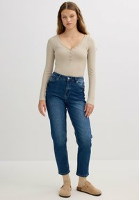 Top de manga larga de color beige claro con detalles de botones, combinado con unos vaqueros de cintura alta de color azul. La modelo lleva unos zapatos de slip-on beige.