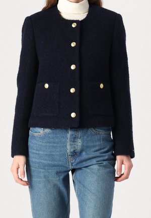 Frau trägt eine marineblaue strukturierte Jacke mit goldenen Knöpfen über einem weißen Rollkragenpullover, kombiniert mit blauen Denim-Jeans, vor einem einfarbigen Hintergrund.