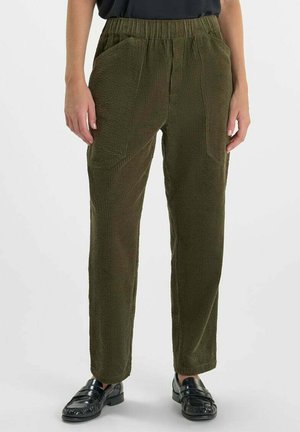 Olijfgroene textuurbroek met een elastische tailleband en frontzakken, gedragen met zwarte loafers door een persoon die tegen een effen achtergrond staat.