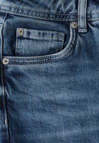 Jeans de mezclilla con un color azul desgastado, un bolsillo frontal con remaches de metal y costuras visibles a lo largo de los bordes.