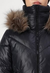 Crna puffer jakna s visokim ovratnikom, koja ima smešasti detalj od smešnog krzna na kapuljači, sjajnu teksturu i dijagonalni kviltani uzorak na tkanini.