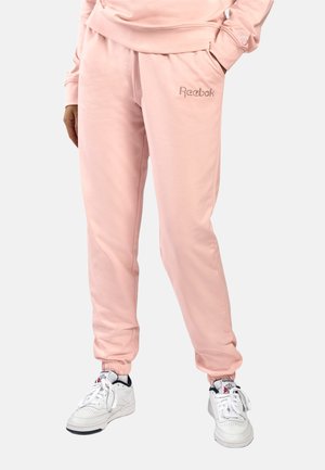 Persoon die lichtroze Reebok joggingbroek en witte Reebok sneakers draagt, staand met handen in de zakken tegen een effen achtergrond.