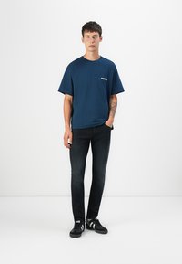 Marineblå bomulds T-shirt med korte ærmer og lige snit, der har et lille grafisk motiv på brystet, parret med mørke jeans og sorte sneakers.