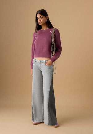 Maglione viola lavorato a maglia con motivo a costine, abbinato a jeans grigi a gamba larga. L'outfit presenta bottoni decorativi e una piccola borsa.