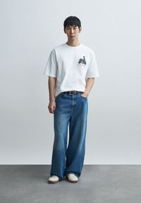 Νέος άνδρας που φοράει λευκό μπλουζάκι με γραφικά, φαρδιά μπλε τζιν, λευκά αθλητικά παπούτσια, στέκεται μπροστά σε γκρι απλό τοίχο με το ένα χέρι στην τσέπη.