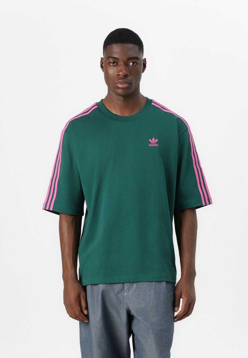 Zielona bawełniana koszulka z różowymi trzema paseczkami na ramionach i małym różowym logo adidas na piersi. Krótkie rękawy i luźny krój.