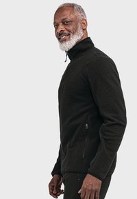 Dunkle, strukturierte Zip-Jacke mit hohem Kragen, ausgestattet mit zwei Seitentaschen und einem subtilen Karomuster. Das Material wirkt warm und strapazierfähig.