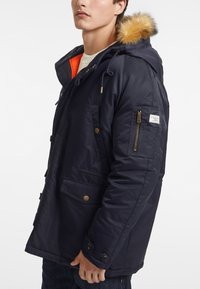 Chaqueta con capucha de color azul marino, con ribete de pelo sintético, bolsillo con cremallera, múltiples bolsillos frontales y un forro naranja. Tela resistente al agua y cierres a presión.