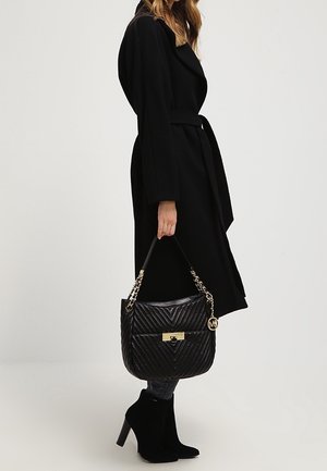 Femme dans un manteau noir ceinturé et des bottines à talons, tenant un sac à main noir matelassé avec une chaîne dorée et un fermoir.