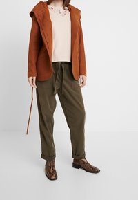 Rdzawoczerwony długi kardigan z kapturem, zestawiony z jasnoróżową bluzką z koronkowym wykończeniem, zielonymi spodniami cargo i wzorzystymi brązowymi botkami za kostkę.