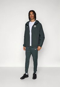 Tumši zaļš Nike sporta kostīms ar rāvējslēdzēja jaku, atbilstošām sporta biksēm, baltu T-kreklu un melnām sporta kurpēm. Uz krūtīm ir logotips.